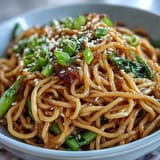 A steaming bowl of Asian ginger scallion noodles with sautéed greens, garnished with sesame seeds and fresh chili for vibrant flavor.